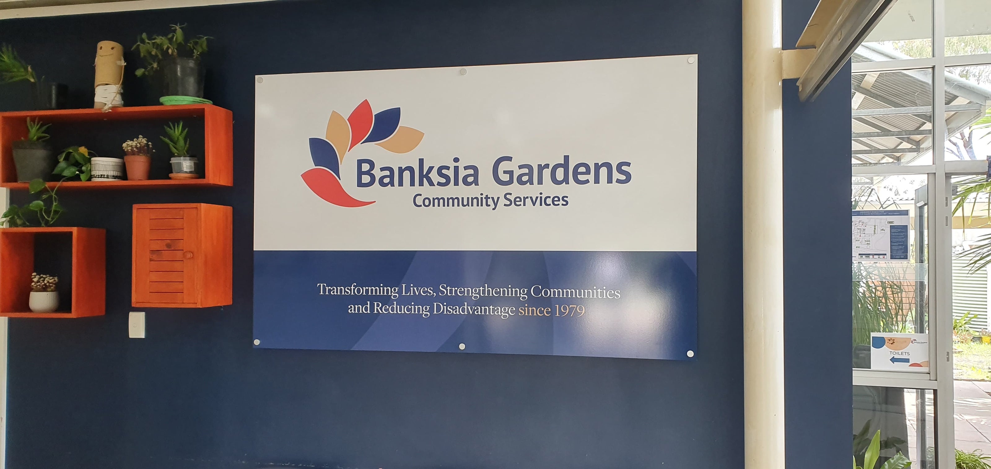 Banksia Gardens Community Services sign on a blue wall with shelves and plants in the background.