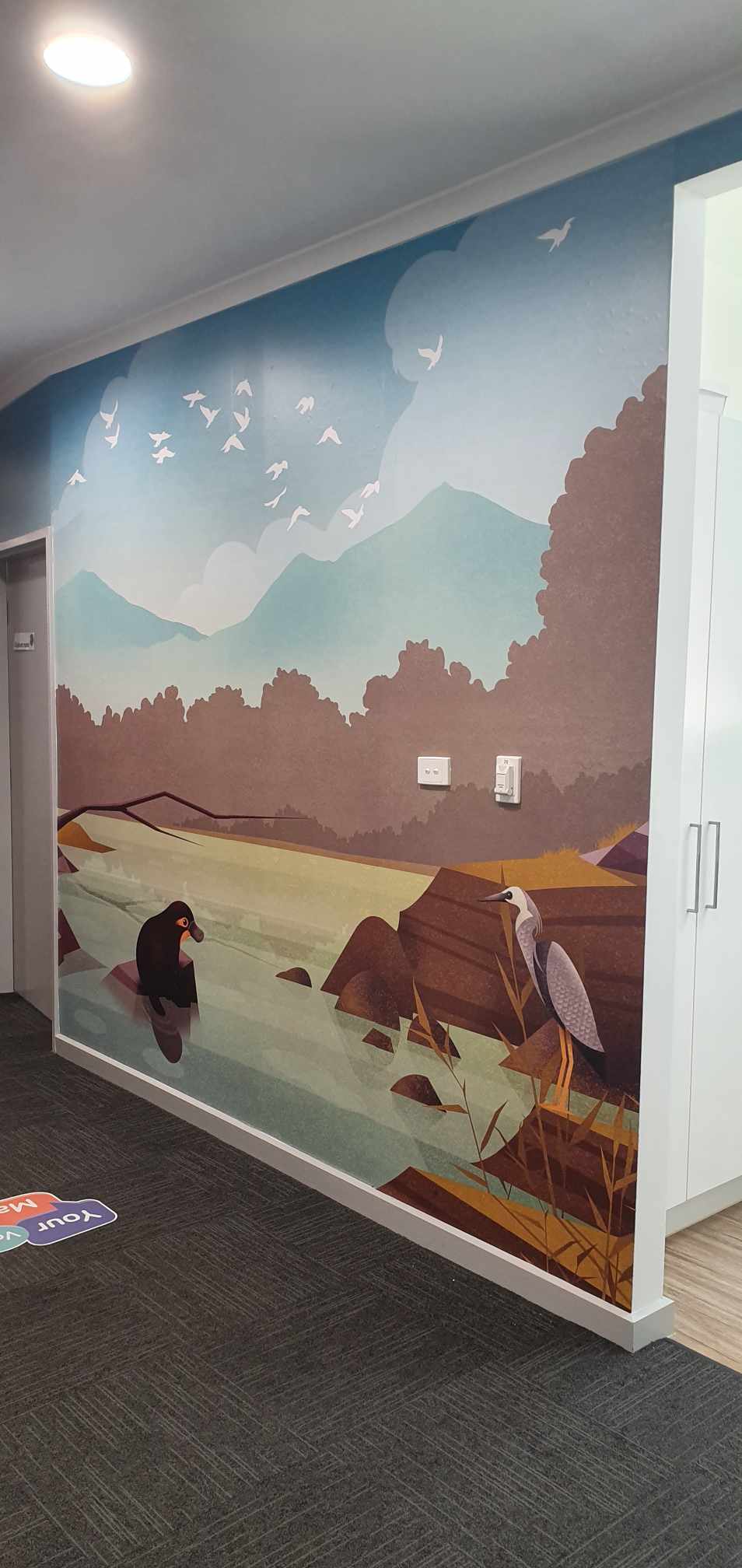 Wall mural of a landscape with mountains, trees, and people by a lake.