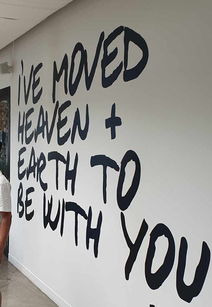 Person standing next to a wall with large black text and a name.