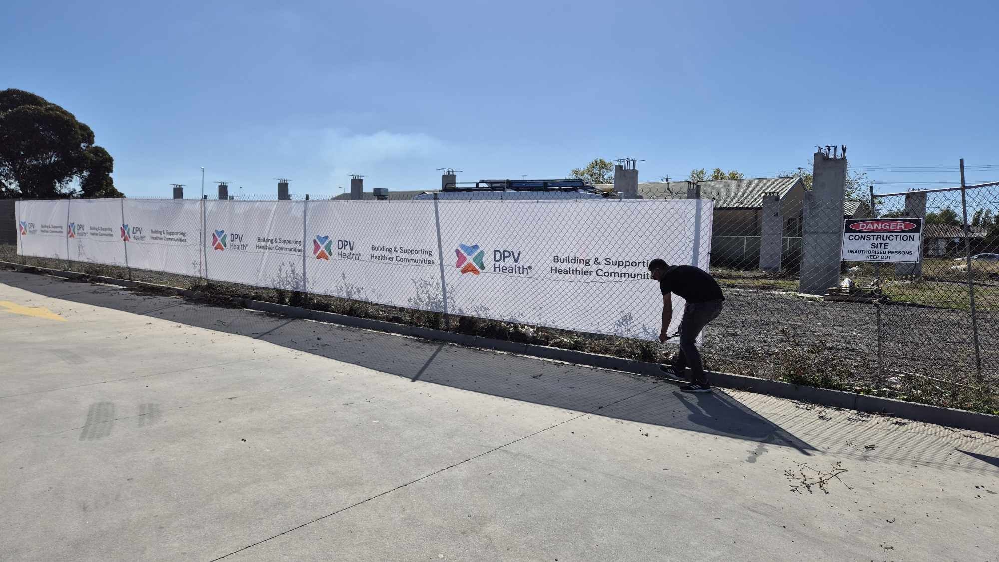 Person standing next to a long white banner with multiple logos on a fence, with a clear blue sky in the background.